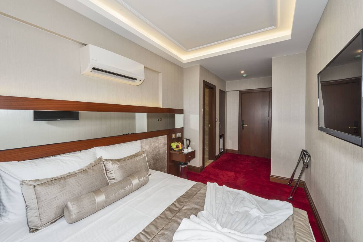 A hotel room with a bed, a bedside table with a lamp, an air conditioning unit, and a red carpet.
