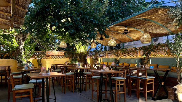 An outdoor dining area with wooden tables and chairs, covered by thatched umbrellas, and surrounded by lush greenery. - Hotel Kostis (Skiathos)