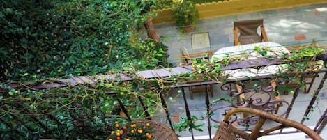A balcony with wicker furniture, a potted plant with yellow flowers, and a view of a building with a tiled roof.