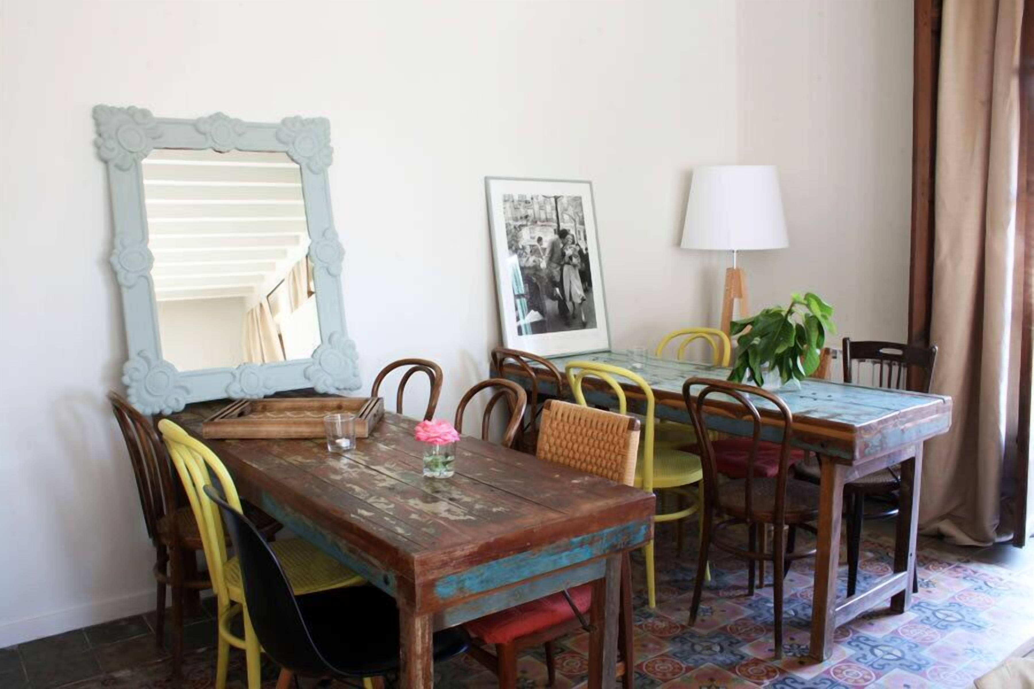 A dining area with a rustic wooden table, colorful chairs, a large mirror, a framed photograph, a lamp, and a potted plant.