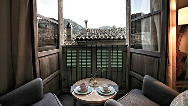 Un rincón acogedor con dos sillones, una mesita redonda con dos tazas y platillos, y una vista de un tejado tradicional con tejas a través de una ventana.