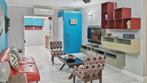 Ein Wohnzimmer mit einem roten Sofa, gemusterten Sesseln, einem Couchtisch, einem Fernseher und einem Bücherregal.