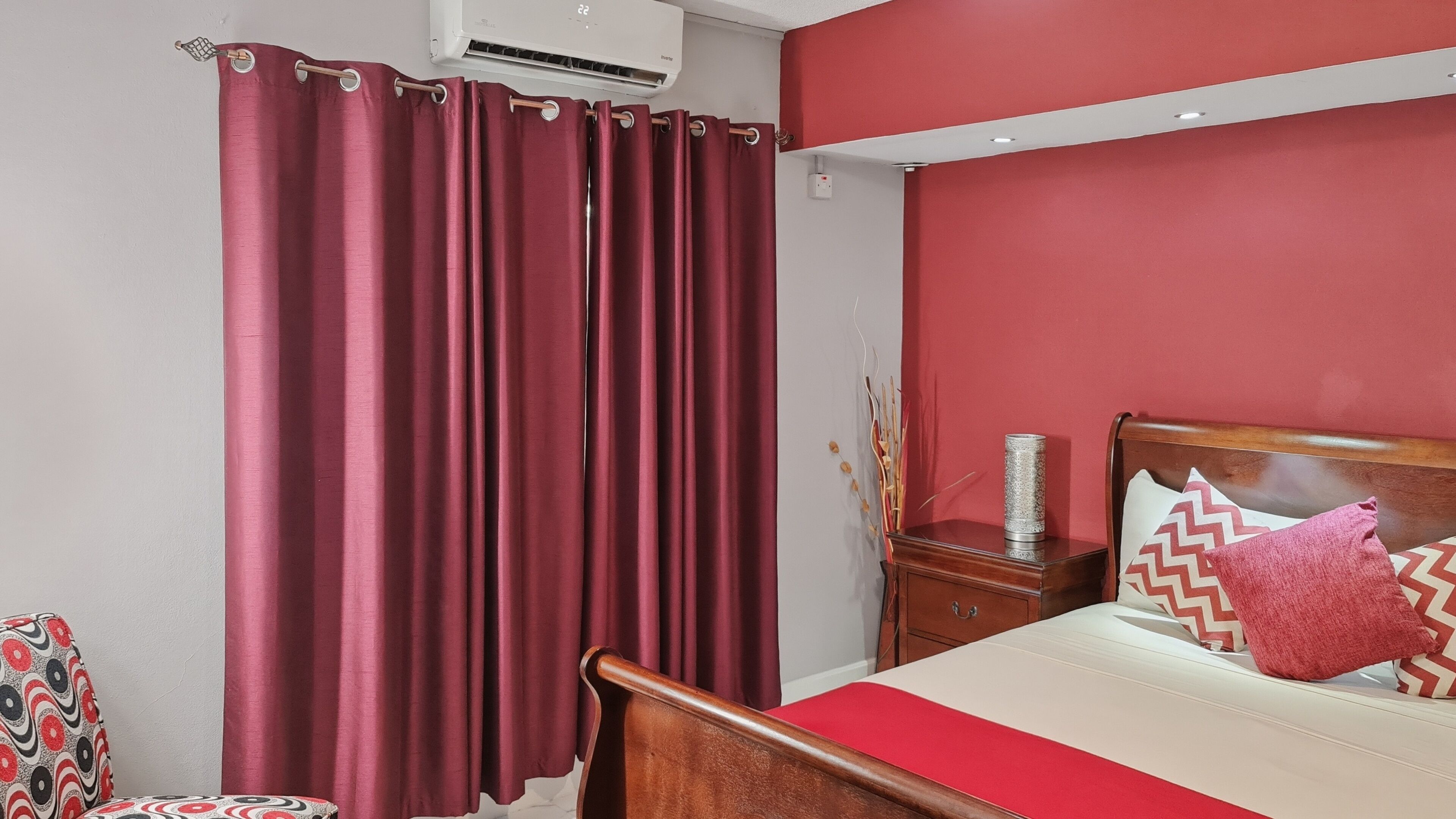 A bedroom with a bed, a nightstand, a red wall, and a window with curtains.