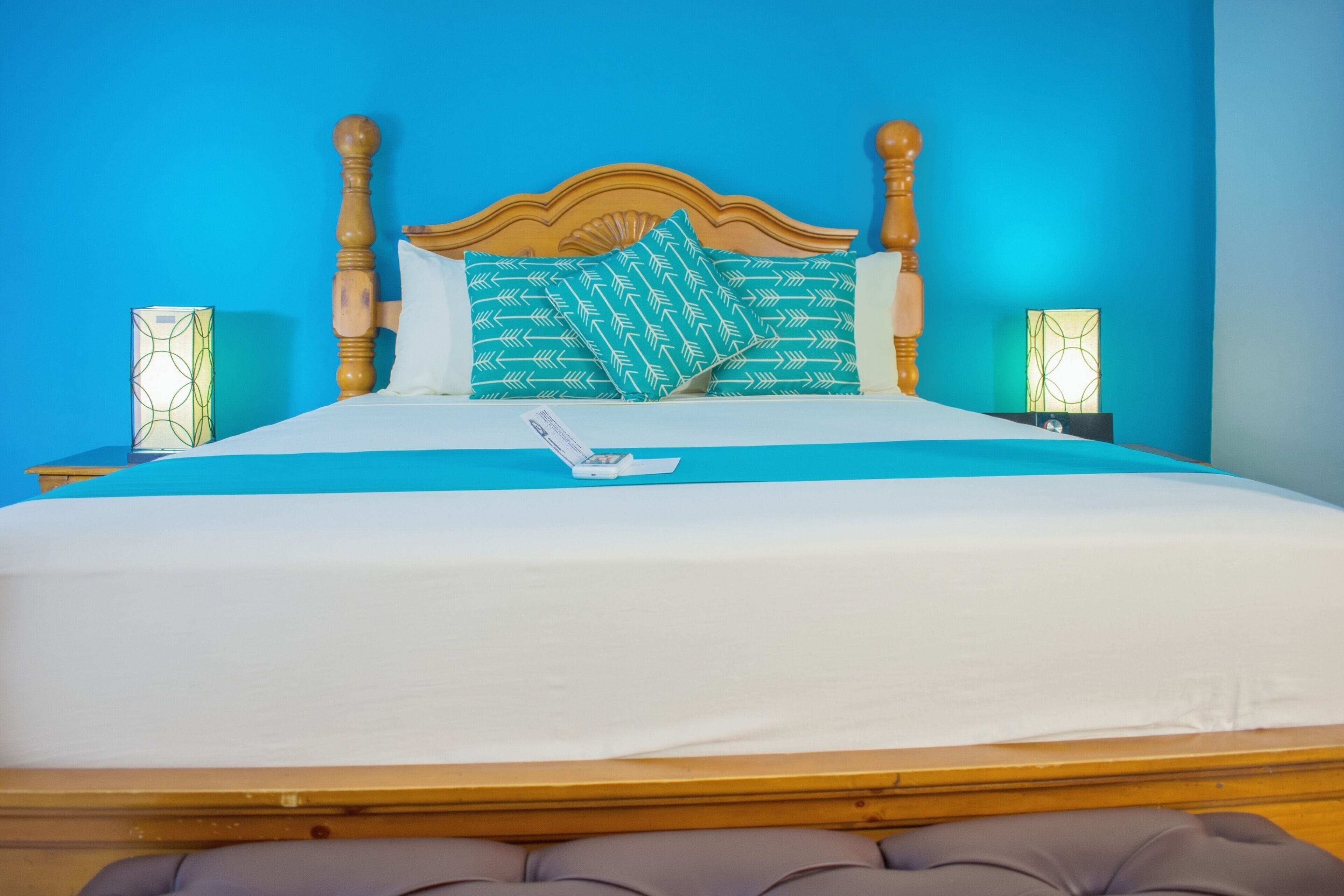 Een bed met een houten hoofdbord, een blauw dekbed en twee kussens met een bladmotief. Er staan twee nachtkastjes met lampen bij.