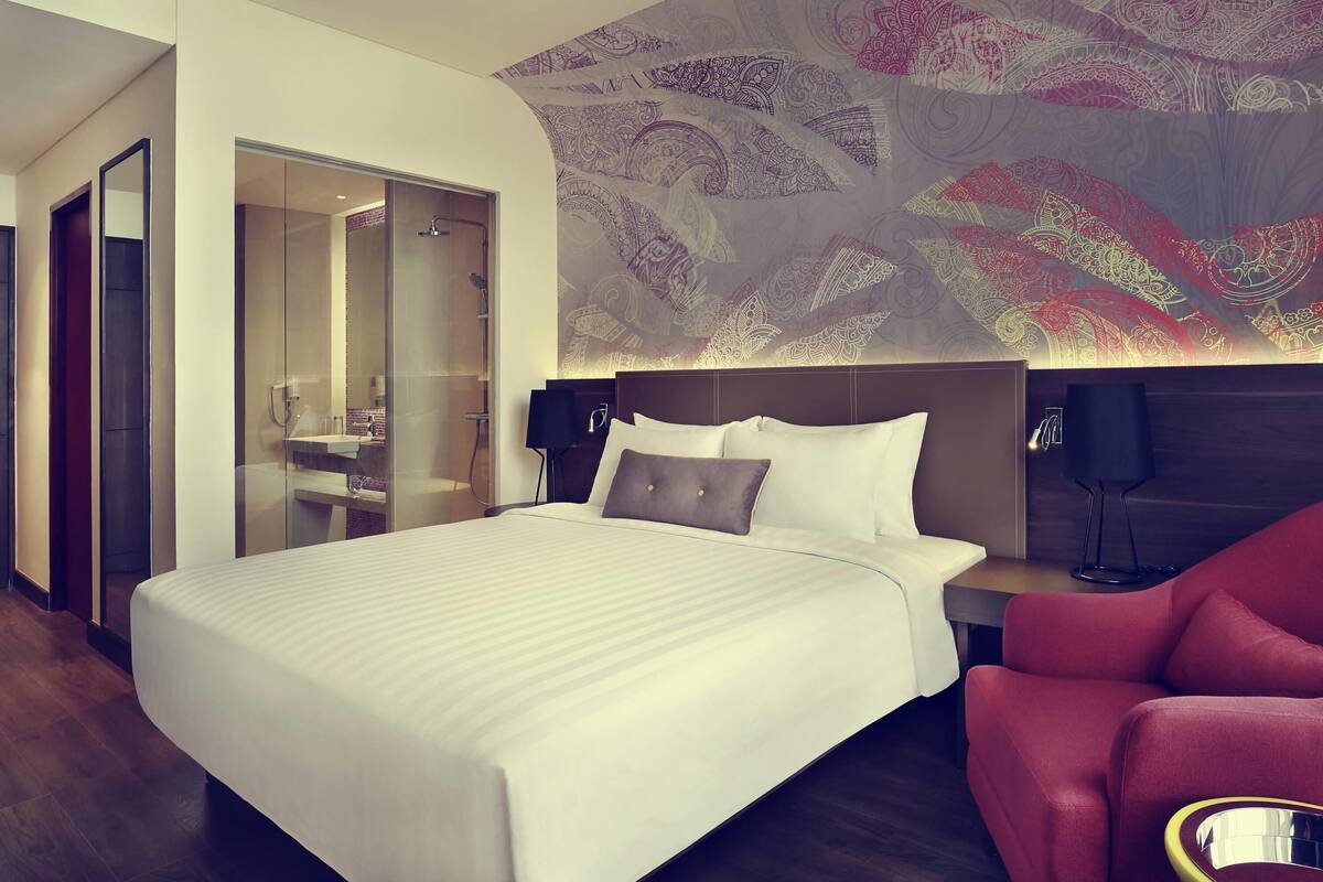 A hotel room with a bed, a red armchair, a nightstand, a wall-mounted lamp, and a glass-enclosed shower area.