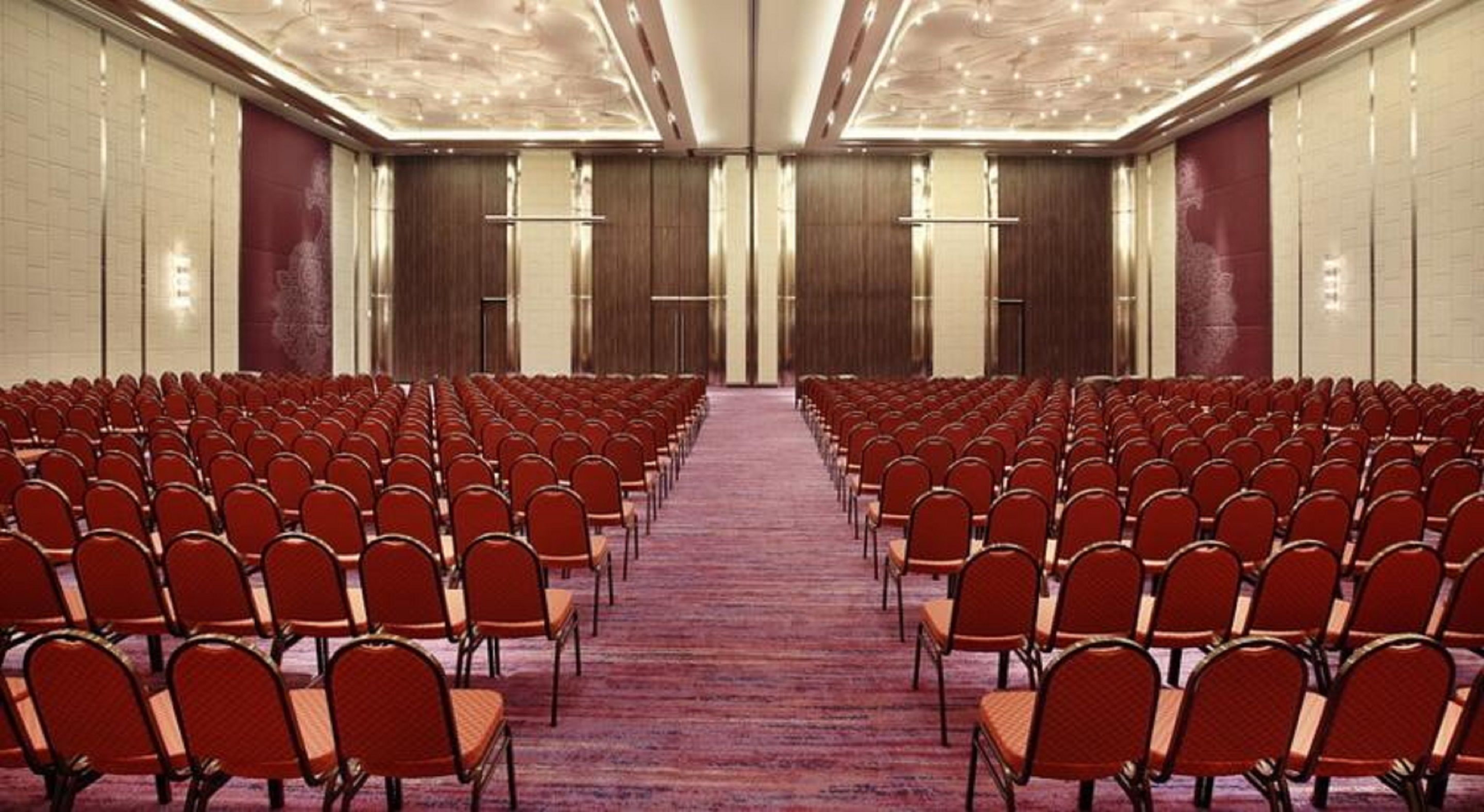 Une salle de conférence avec des rangées de chaises rouges, un sol recouvert de moquette et un plafond haut doté d’un éclairage encastré.