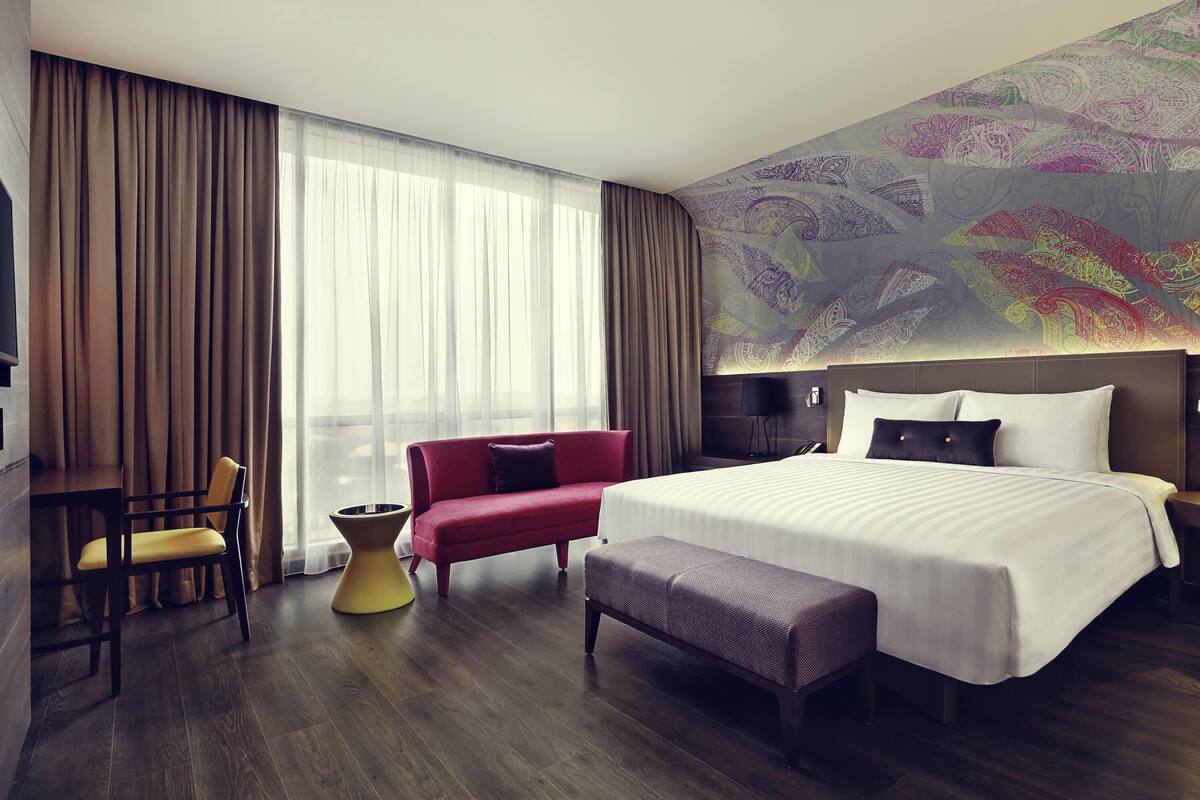 A hotel room with a large bed, a red sofa, a yellow chair, a small table, and a large window with curtains.