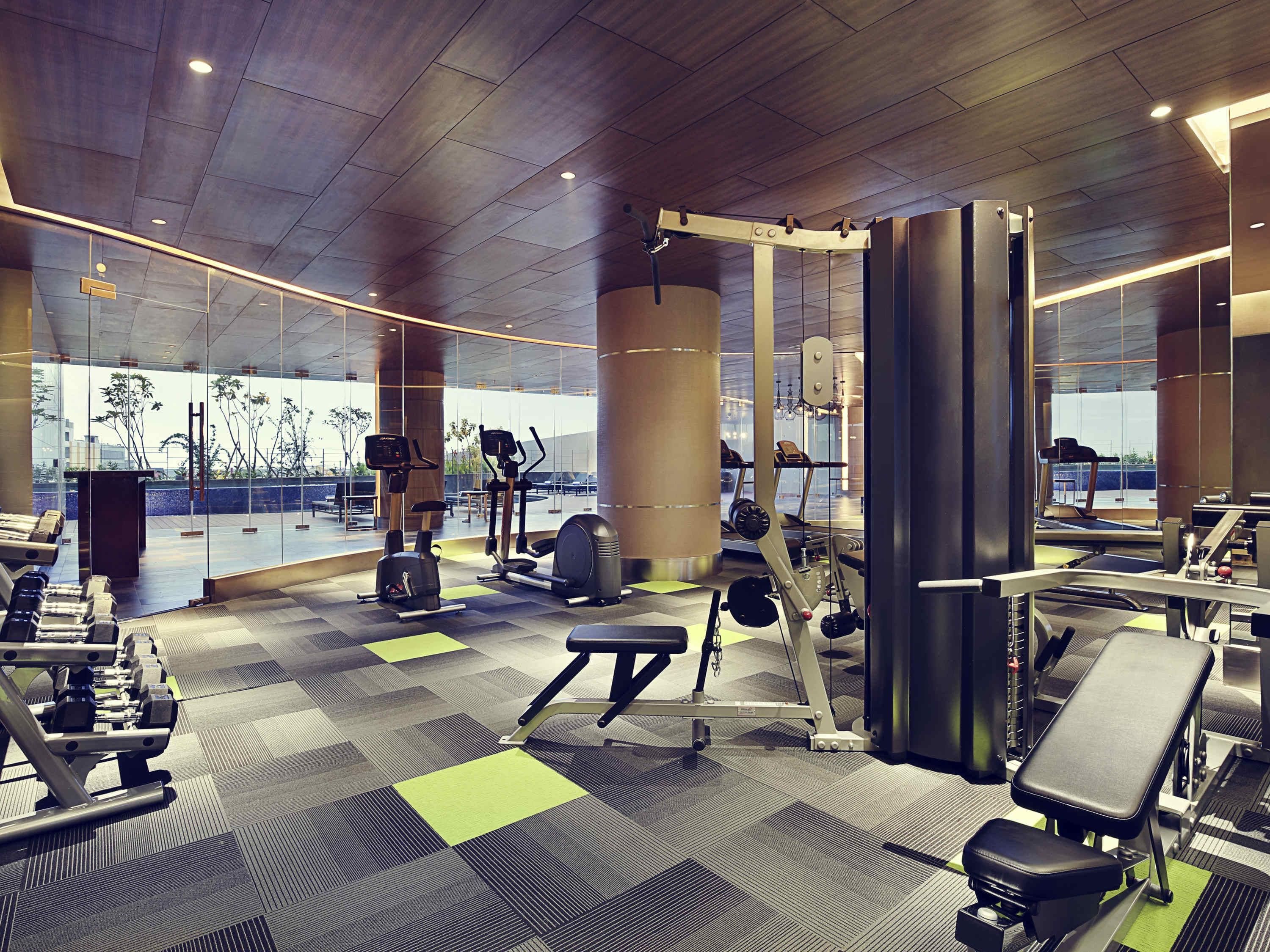 A modern gym with various exercise equipment, large windows, and a patterned floor.