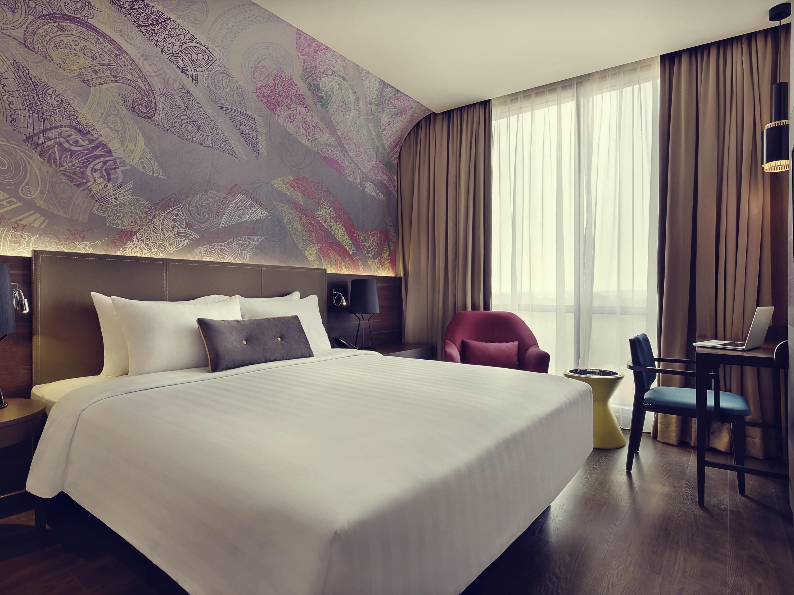 A hotel room with a large bed, a desk with a laptop, a chair, and a decorative wall.