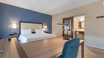 Habitación de hotel con cama, mesitas de noche, escritorio, silla, espejo y un baño visible a través de una puerta abierta.