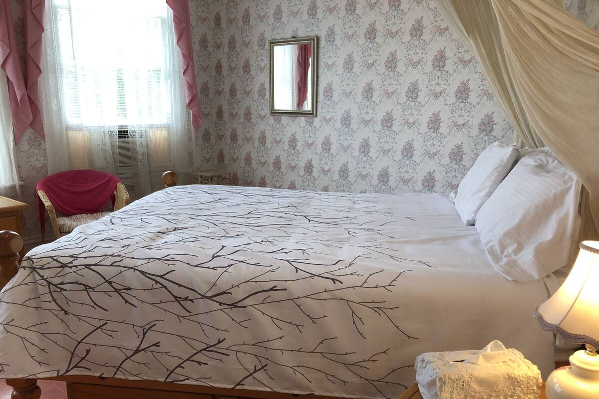 Een net opgemaakt bed met een sprei met een wit en zwart takkenpatroon, een houten nachtkastje met een lamp, een stoel met een roze plaid, en bloemenbehang.