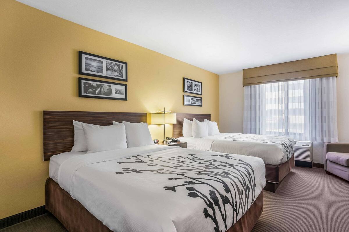 A hotel room with two beds, a nightstand, a lamp, and a window with curtains.