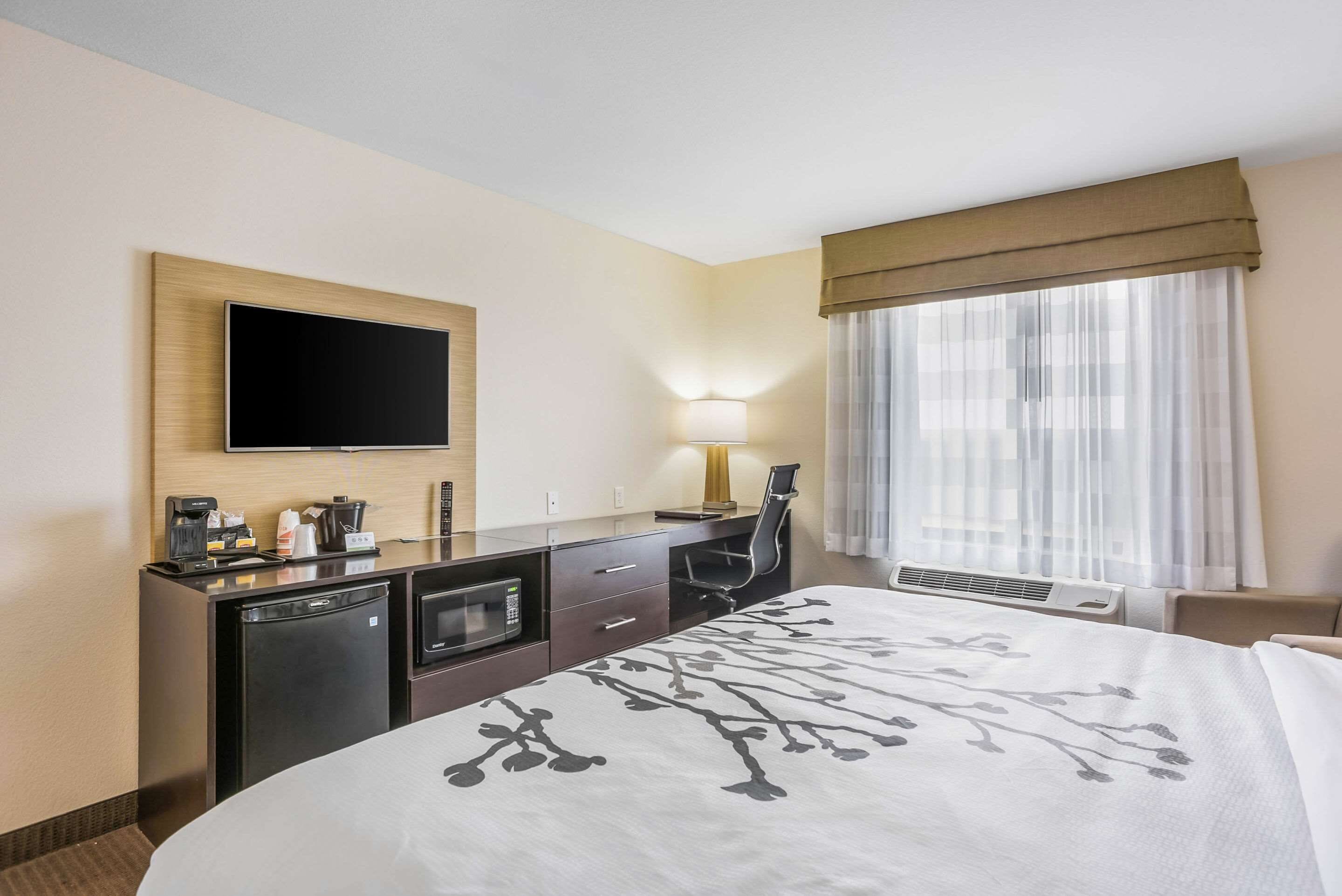 Ein Hotelzimmer mit Bett, Schreibtisch mit Flachbildfernseher, Kaffeemaschine, Mikrowelle, Lampe und Fenster mit durchsichtigen Vorhängen.