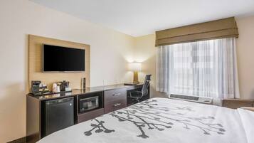Ein Hotelzimmer mit Bett, Schreibtisch mit Flachbildfernseher, Kaffeemaschine, Mikrowelle, Lampe und Fenster mit durchsichtigen Vorhängen.