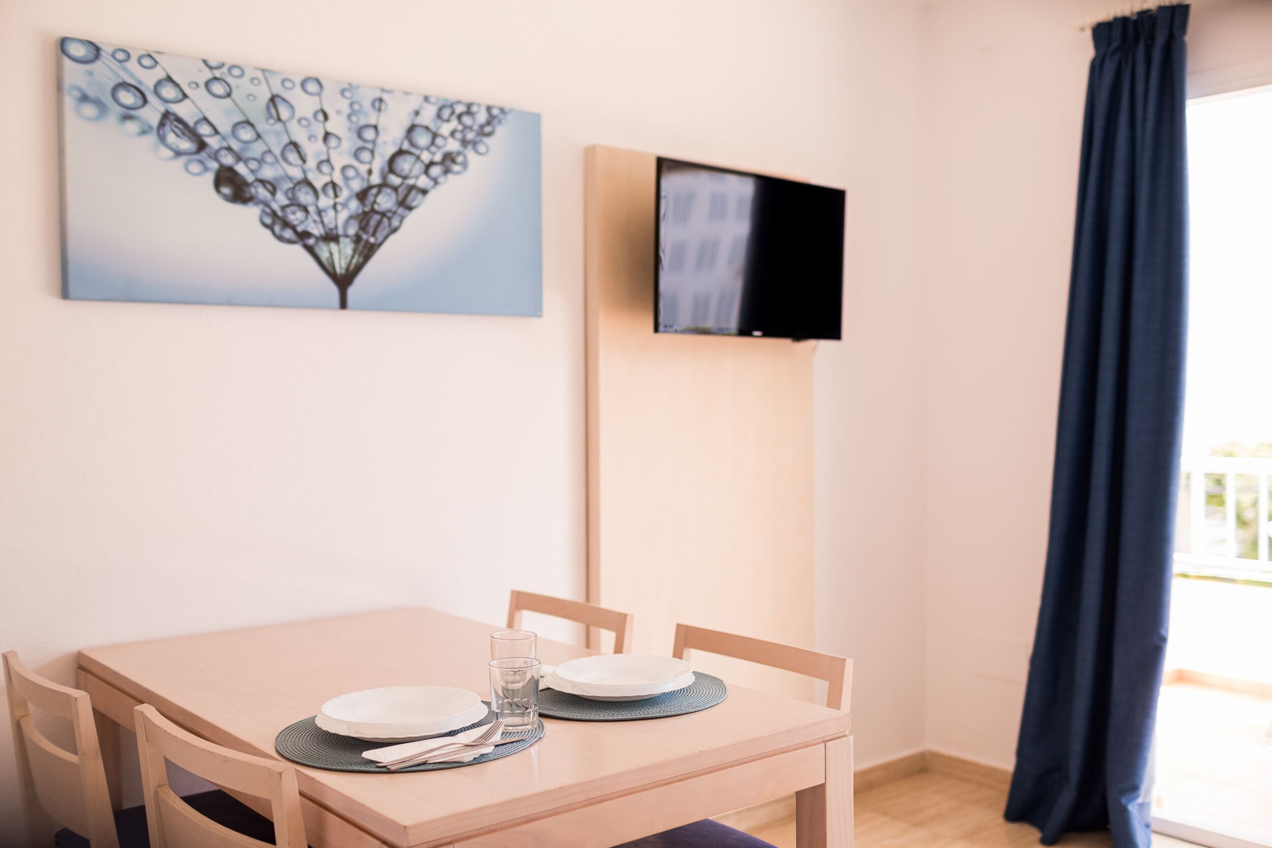 Un comedor con una mesa preparada para dos personas, un televisor de pantalla plana montado en la pared y una ventana con cortina oscura.