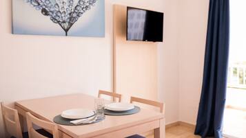 Un comedor con una mesa preparada para dos personas, un televisor de pantalla plana montado en la pared y una ventana con cortina oscura.