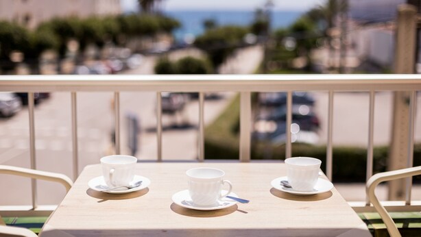 A balcony with two white cups on saucers, a spoon, and a view of the sea and buildings in the background. - Vistamar I - MC Apartaments Ibiza (Ibiza Town)
