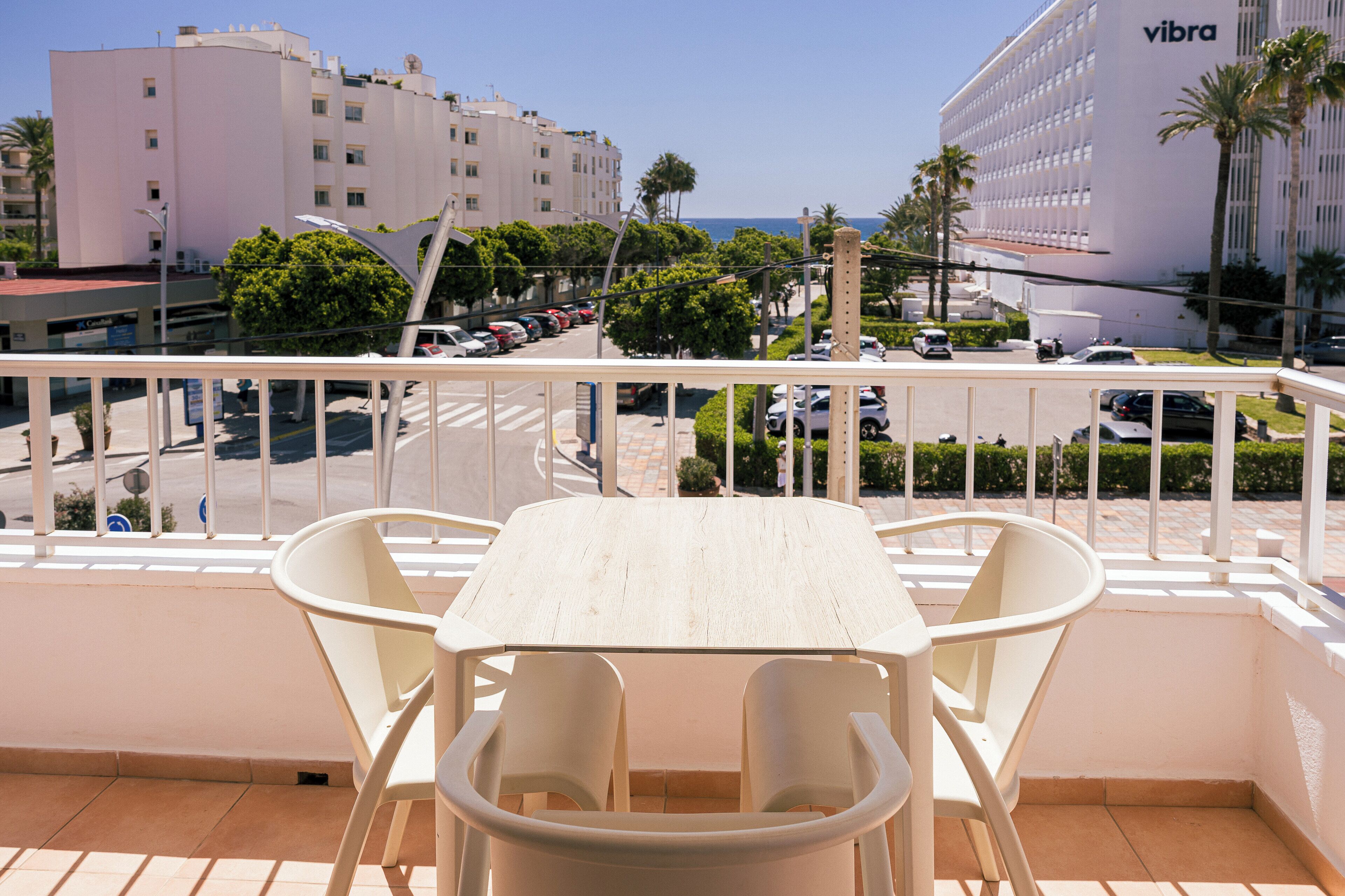 A balcony with a white table and chairs, overlooking a street with parked cars and buildings, including a 'Vibra' hotel.