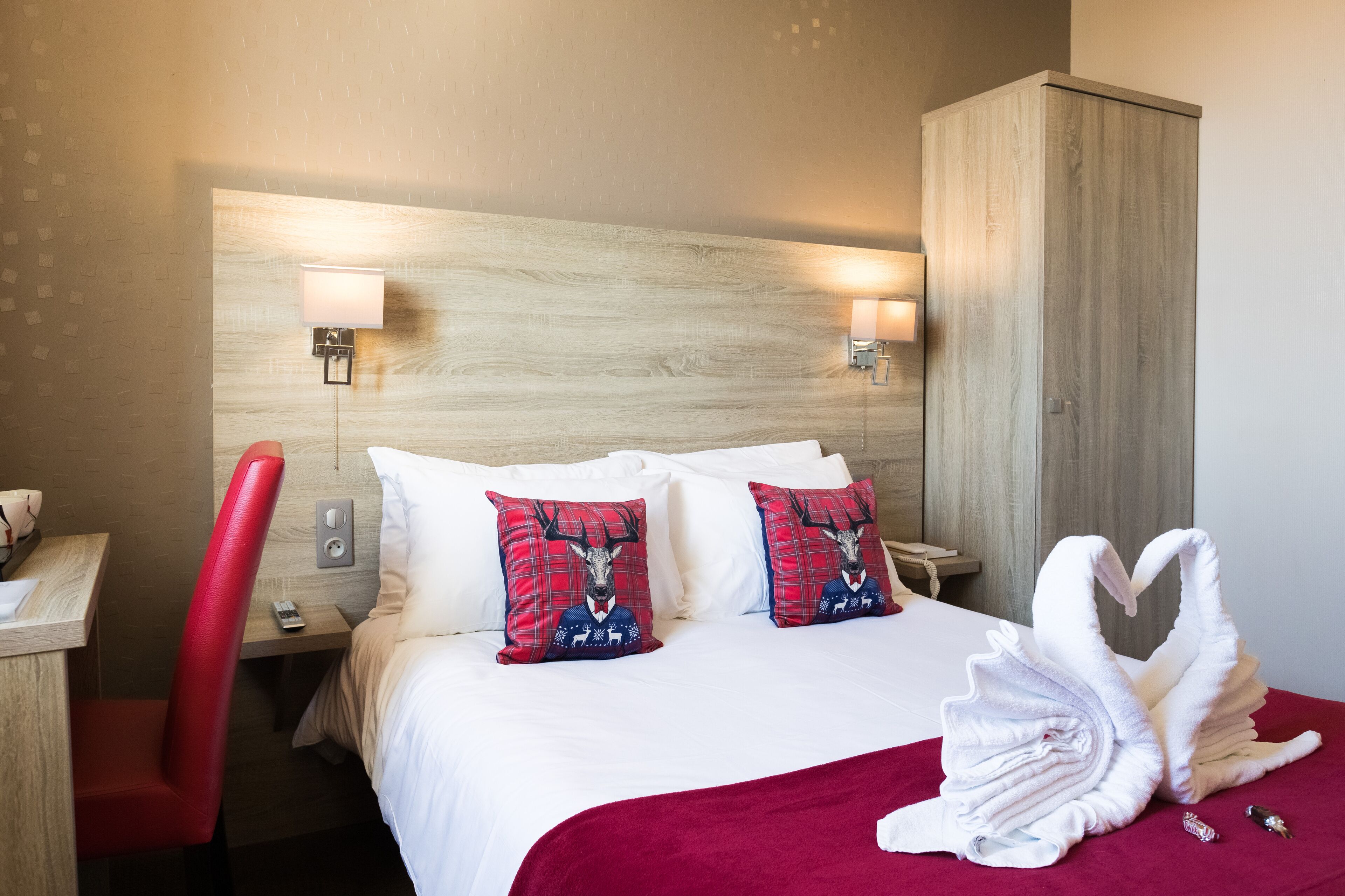 A hotel room with a bed, two pillows with deer designs, a red chair, a wooden wardrobe, and a red blanket.