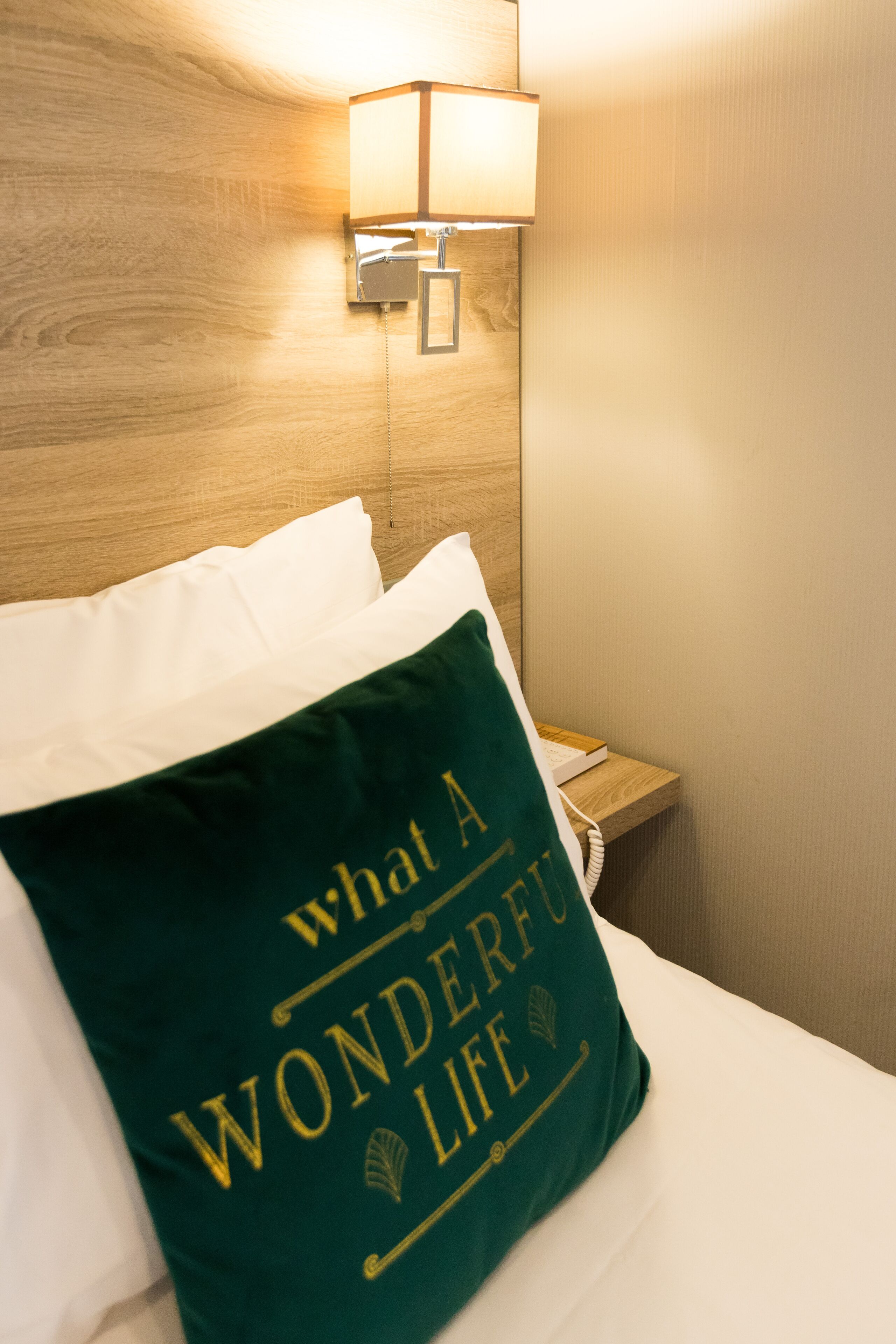 Een net opgemaakt bed met een groen kussen waarop staat 'What a Wonderful Life' en een bedlampje.