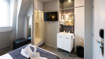 Una habitación de hotel moderna con una cama, ducha, televisor montado en la pared, baño blanco con lavabo y un cubo de basura negro.