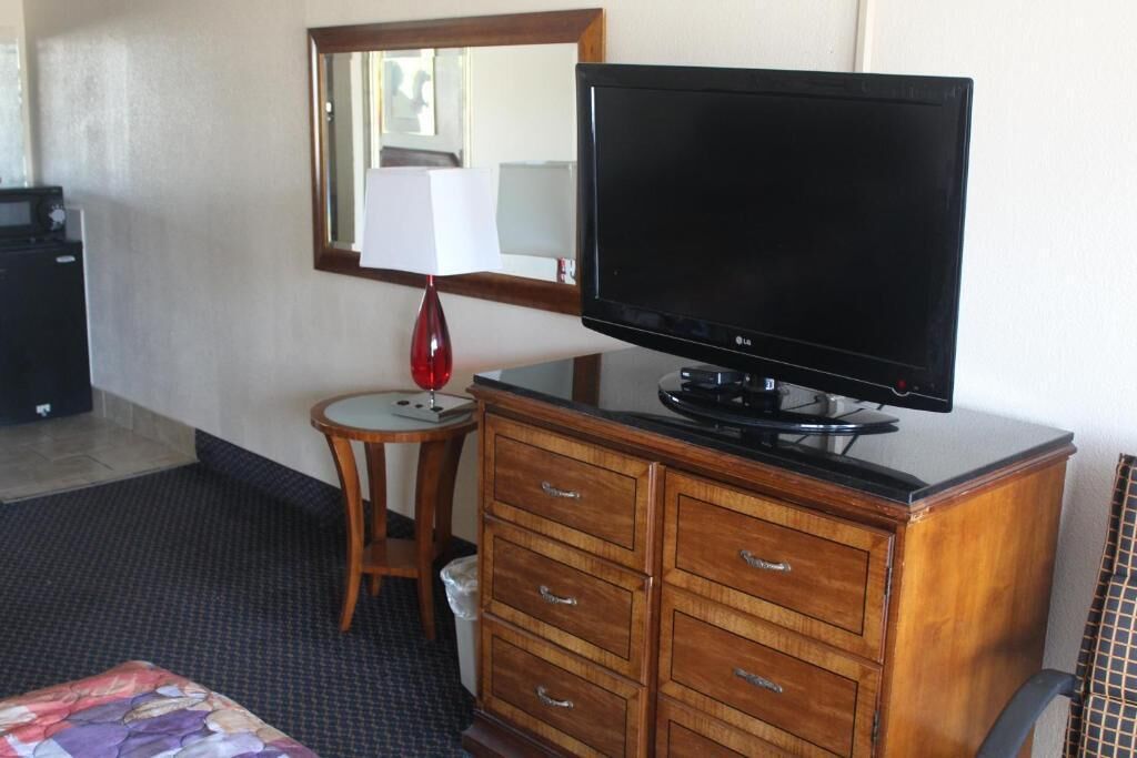 Ein Hotelzimmer mit einem hölzernen Schminktisch, einem Fernseher darauf, einem Spiegel über dem Tisch und einem kleinen runden Tisch mit einer Lampe, sowie einem Bett mit einer farbenfrohen Decke.