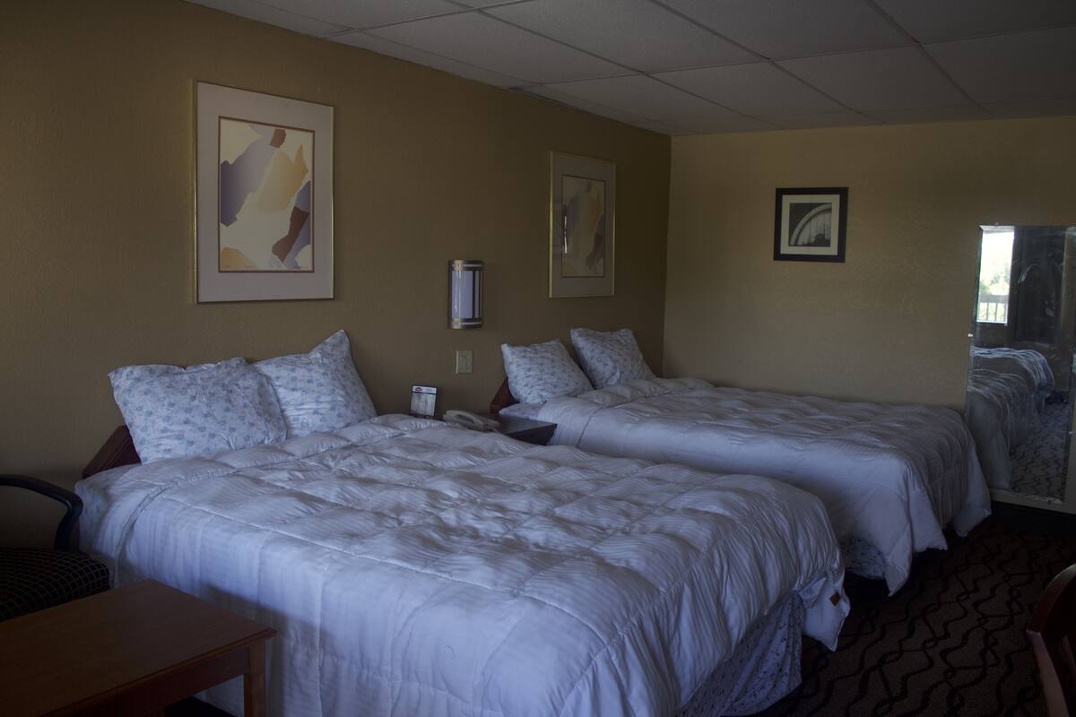 Een hotelkamer met twee bedden, een bureau en een stoel. Er hangen afbeeldingen in lijsten aan de muur en een raam met jaloezieën.