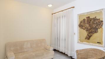 Quarto com sofá bege, cadeira de vime, pintura abstrata grande e janela com cortinas finas.