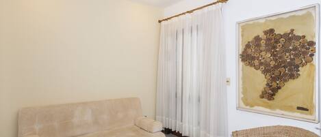 A room with a beige sofa, a wicker chair, a large abstract painting, and a window with sheer curtains.