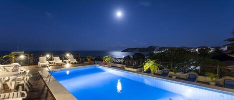 A nighttime pool area with lounge chairs and a view of the sea.