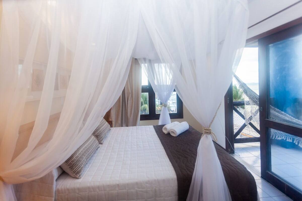A room with a bed, white curtains, and a view of the sea through a window.