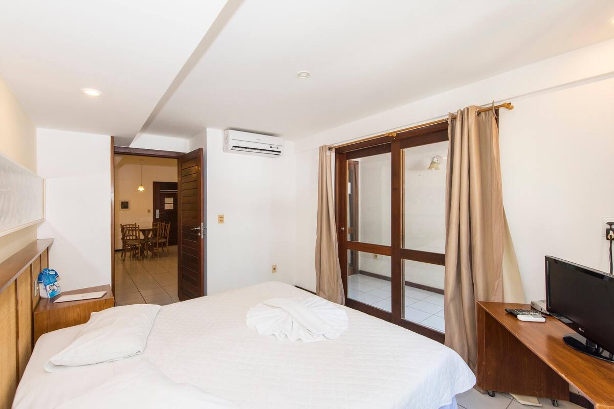 A hotel room with a bed, a desk with a TV, an air conditioning unit, and a sliding glass door to the balcony.
