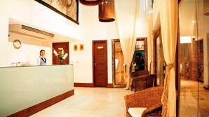 Zona de recepción de un hotel con recepcionista, mostrador, sillas de mimbre y un espejo grande que refleja el interior.