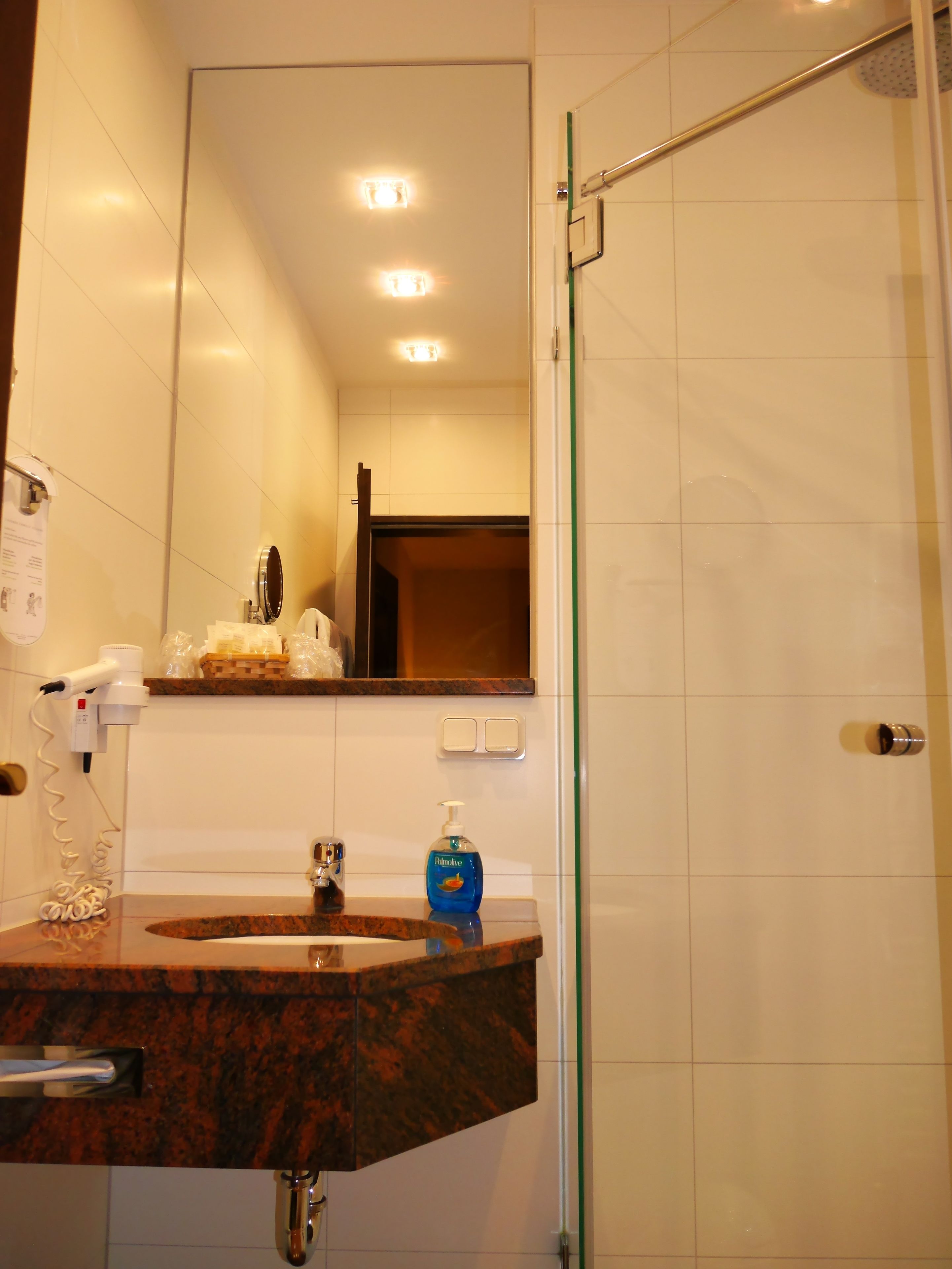 Un baño con lavamanos de mármol, espejo, secador de pelo, dispensador de jabón y puerta de ducha.