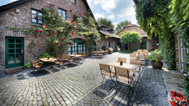 A courtyard with cobblestone flooring, wooden tables and chairs, and brick buildings with green doors and windows. - Landgut Ramshof (Willich)