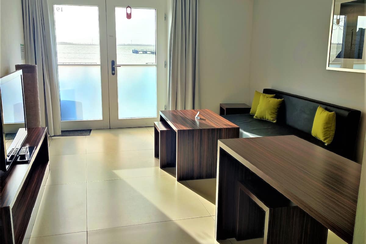 A modern hotel room with a large sliding glass door leading to a balcony, a flat-screen TV, a wooden dining table, a sofa with yellow cushions, and a framed picture on the wall.