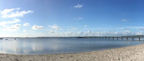 Een rustig strand met een zandkust, een pier die het water in reikt, en enkele boten.