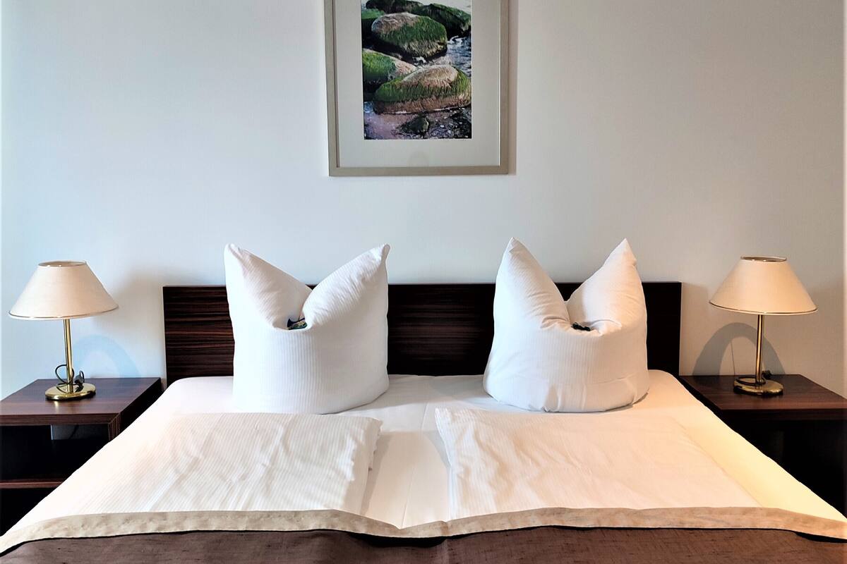A neatly made bed with white linens and two pillows, flanked by two bedside tables with lamps, in a room with a framed landscape painting on the wall.