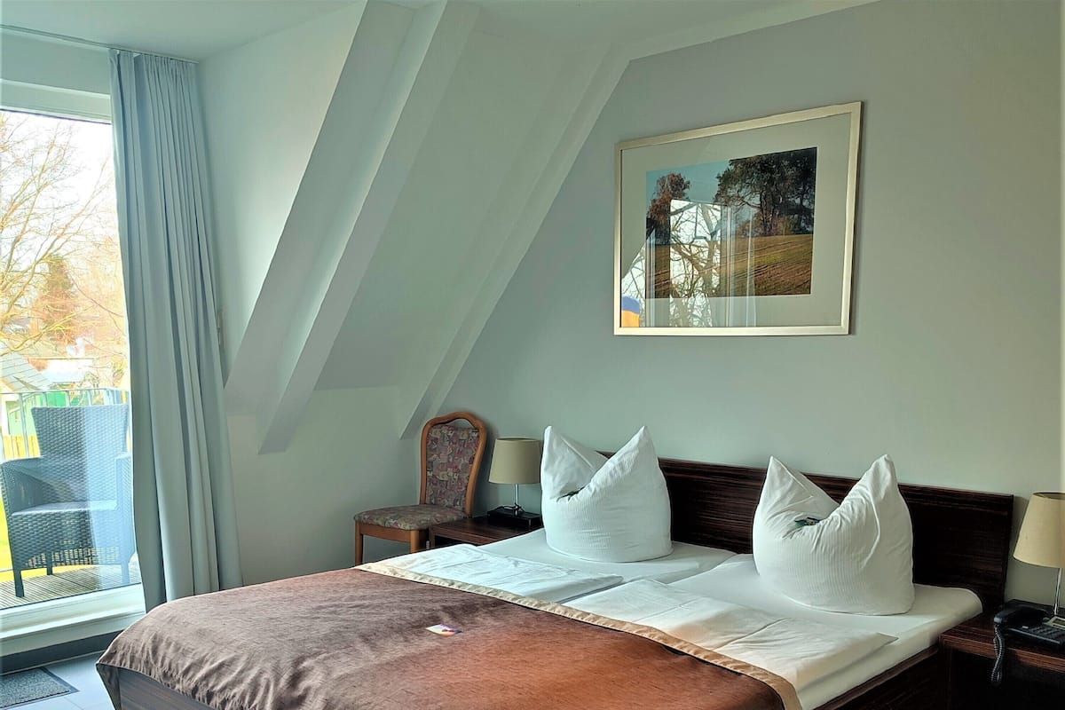 A neatly made bed with a brown cover, a chair, a bedside table with a lamp, and a painting on the wall.