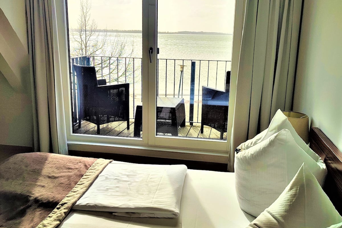 A hotel room with a large window overlooking a deck with chairs and a view of the water.