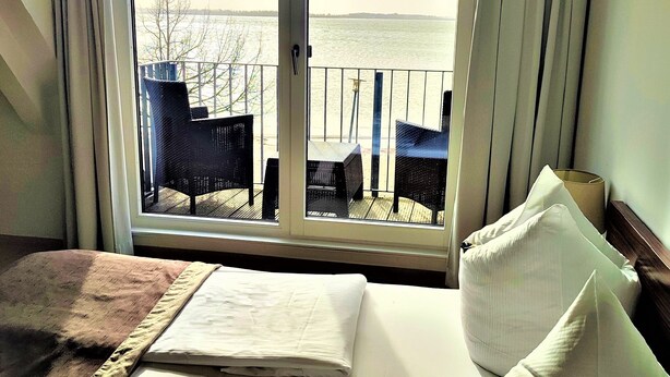 Habitación de hotel con un ventanal grande que da a una terraza con sillas y vistas al agua.
