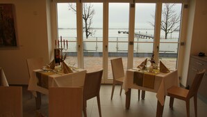 Un comedor con mesas dispuestas para una comida, vista a un cuerpo de agua a través de amplias ventanas y un cuadro en la pared.