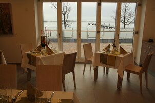 A dining area with tables set for a meal, a view of a body of water through large windows, and a painting on the wall.