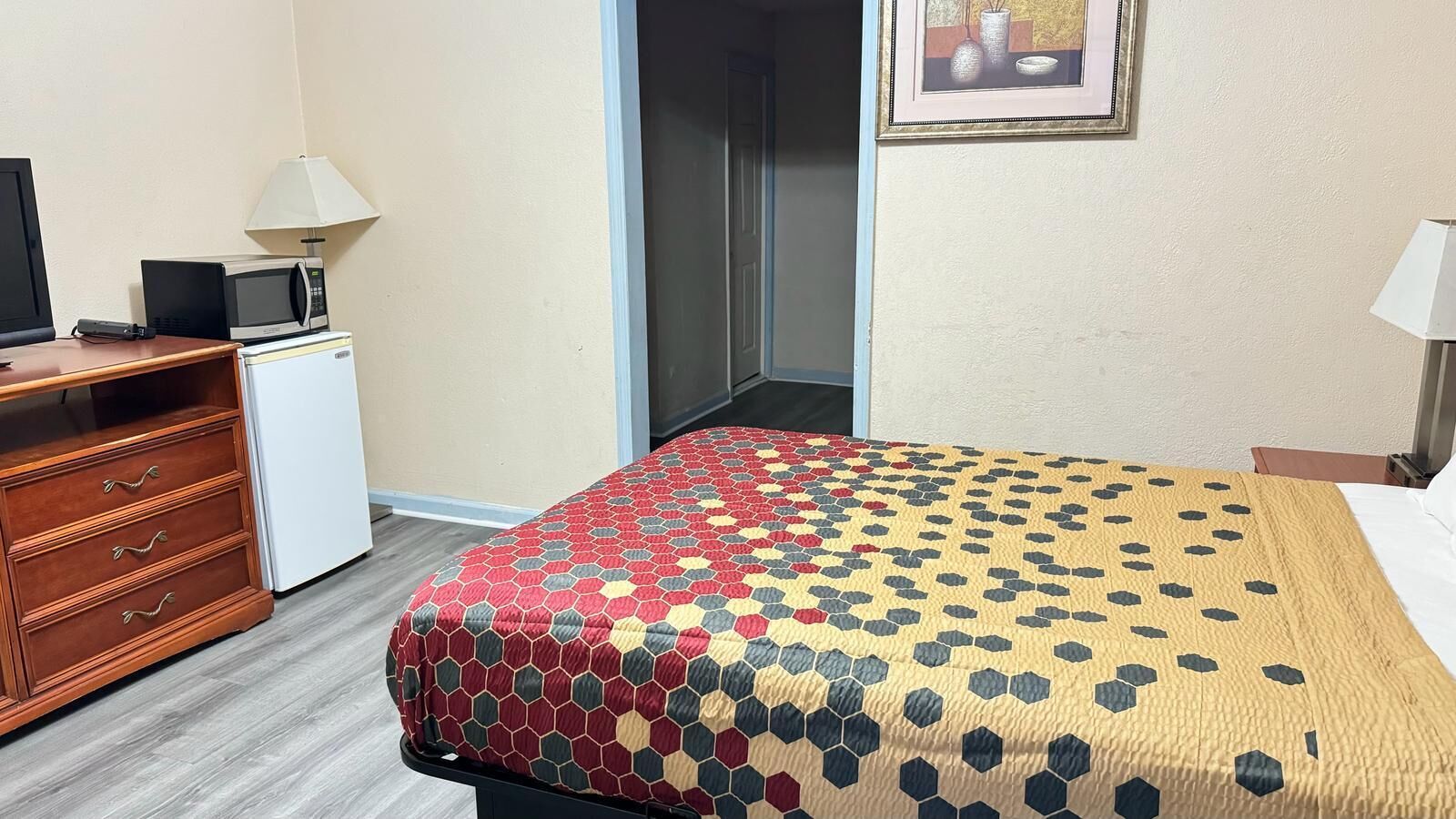 Habitación de hotel con cama, cómoda, microondas, refrigerador pequeño y puerta que da a un pasillo.