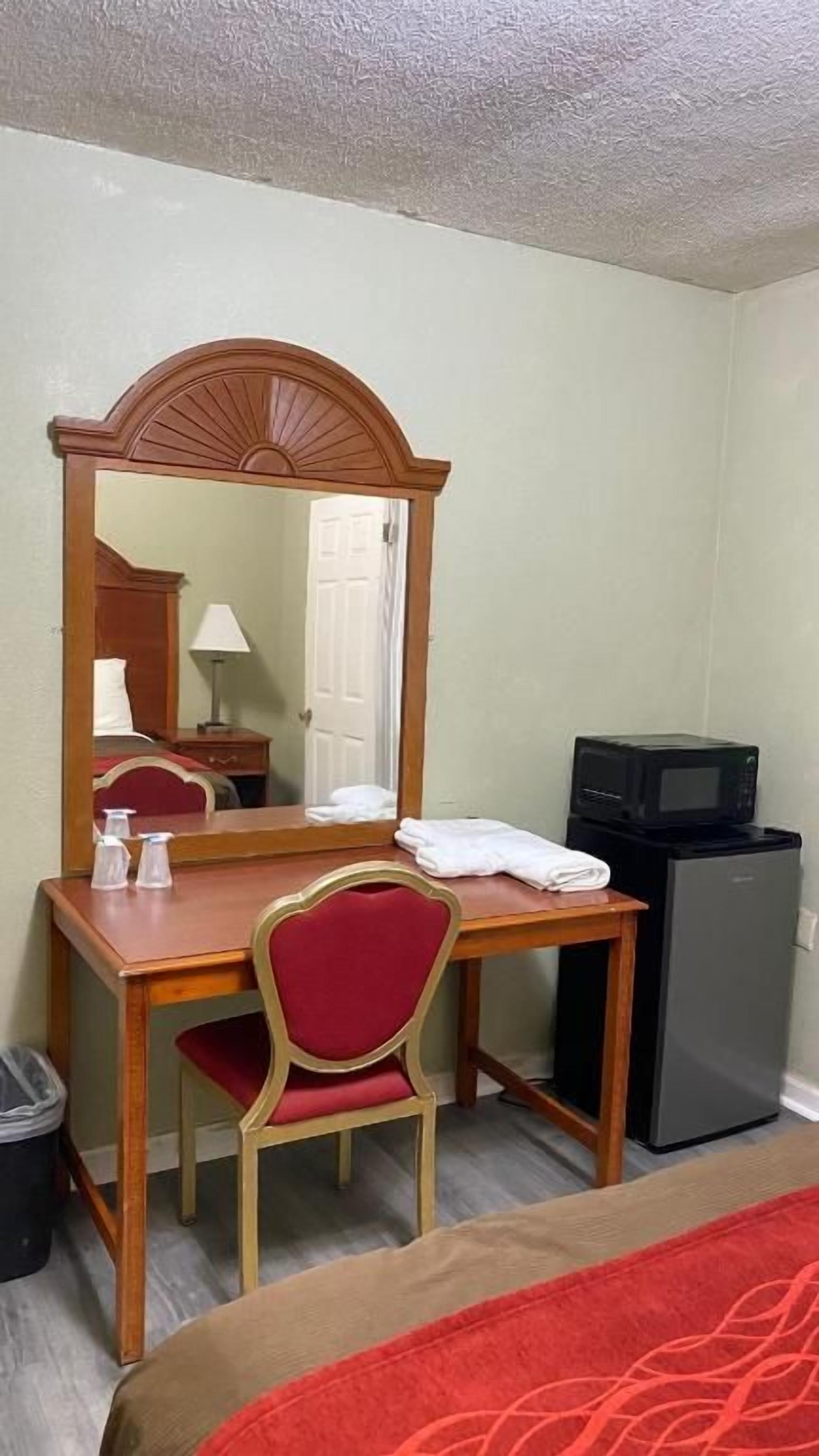 A hotel room with a wooden desk, a chair, a mirror, a bed, a television, and a small refrigerator.
