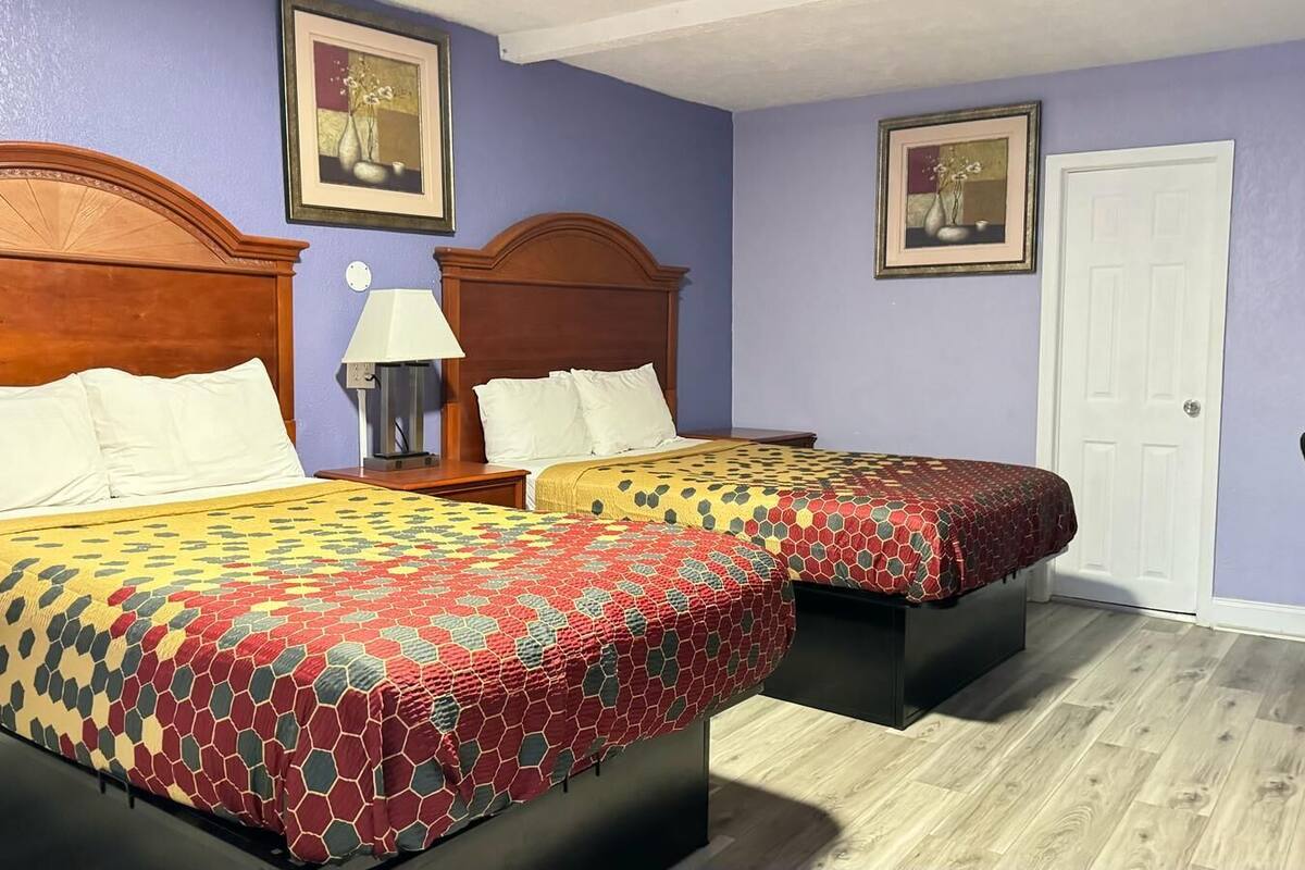 A room with two beds, each with a wooden headboard, a nightstand with a lamp, and a framed picture on the wall.