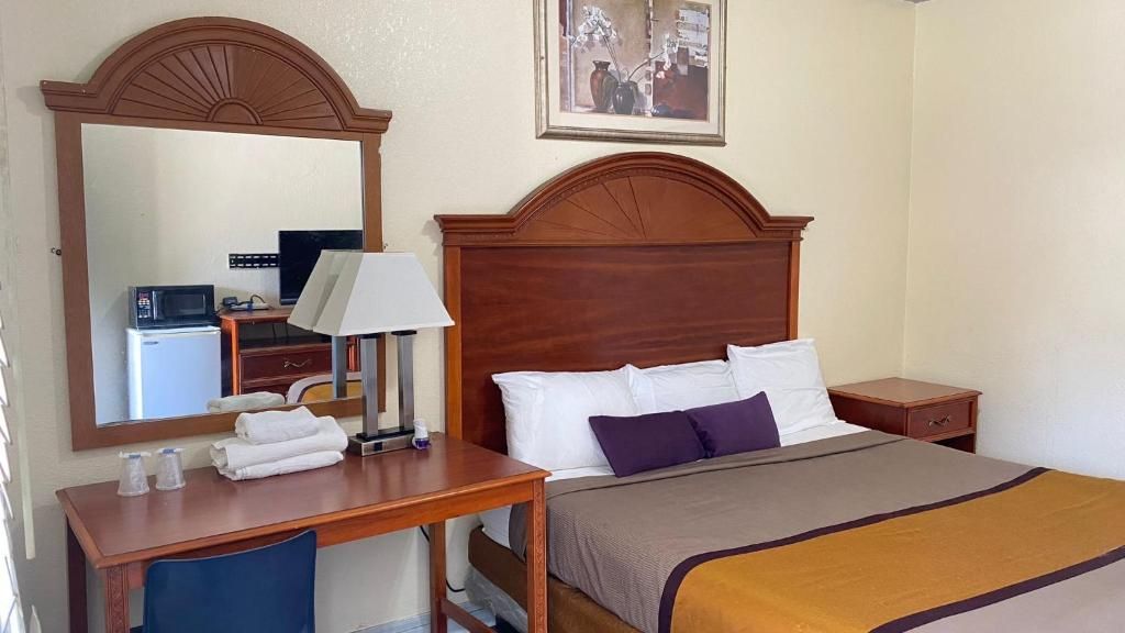 A hotel room with a wooden headboard, a bed with white and purple bedding, a desk with a lamp, and a mirror reflecting a kitchenette.