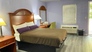 A hotel room with a bed, bedside table, lamp, chair, and an air conditioning unit.
