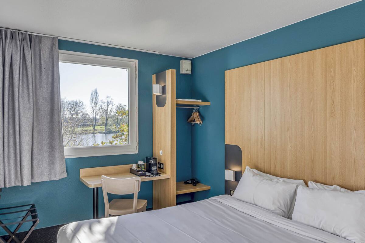 Une chambre d’hôtel avec un lit, un bureau, une chaise, une fenêtre avec des rideaux et une vue sur les arbres.