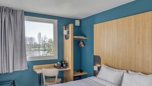 A hotel room with a bed, a desk, a chair, a window with curtains, and a view of trees.